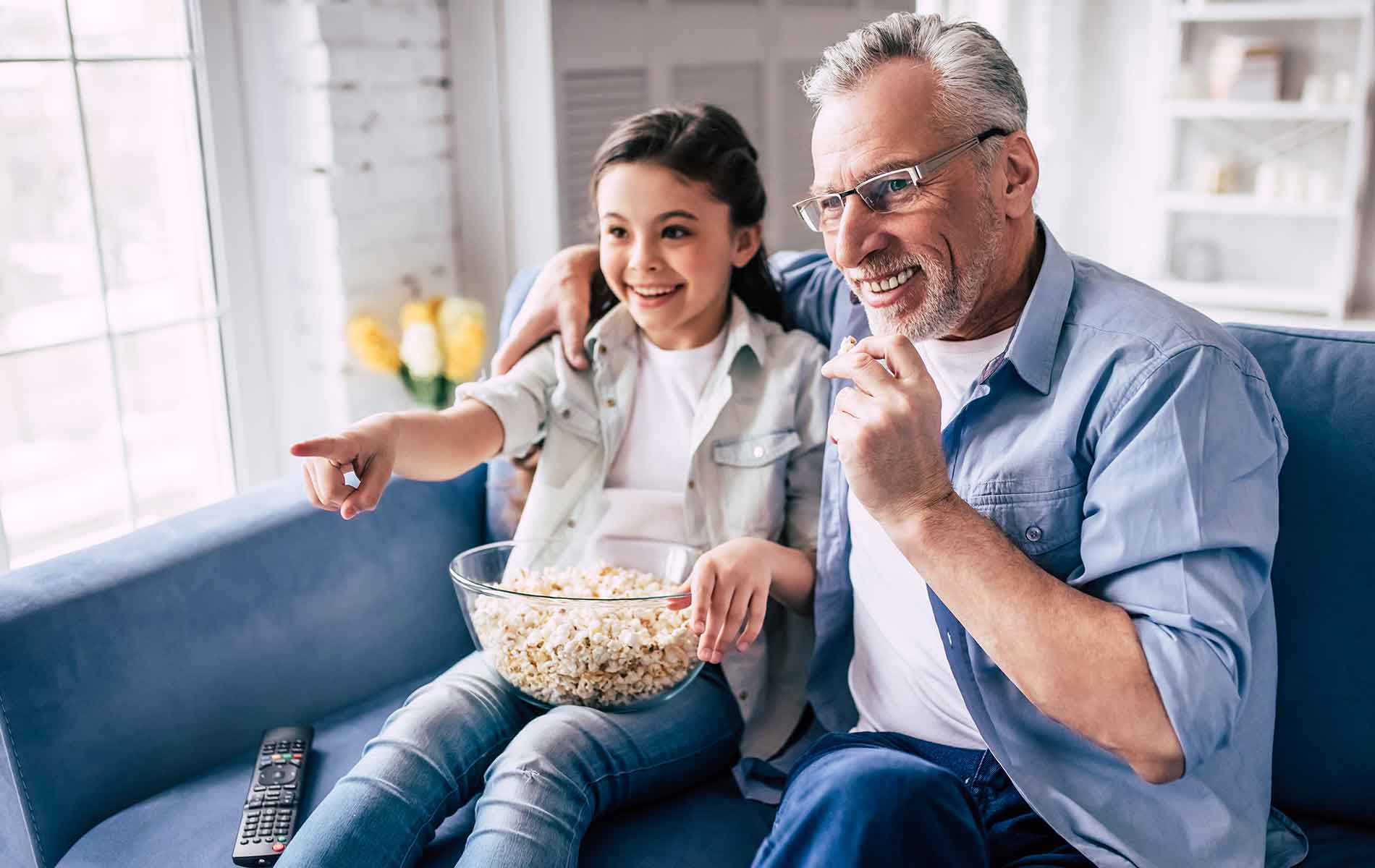 Grand dad with his grand daughter sharing bowl of popcorn sitting on the sofa 